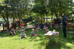 W parku na terenie przedszkola w Wieliczce, funkcjonariusz KPP w Wieliczce przeprowadza pogadankę z przedszkolakami.