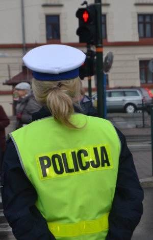 policjantka ruchu drogowego obserwująca  przejście dla pieszych z sygnalizacją świetlną. W tle widoczny jest jasny budynek oraz stojące samochody oraz kobieta