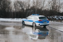 Fotografia przedstawia radiowóz policyjny marki Kia Ceed o nr rej HPGC-143 i numerze taktycznym G484 podczas wykonywania ćwiczenia na płycie poślizgowej. Widoczny od tyłu samochód znajduje się w poślizgu nadsterownym. Na podłożu widać cienką taflę wody.