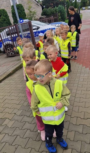 Przedszkolaki poruszające się chodnikiem. W trle radiowóz policyjny