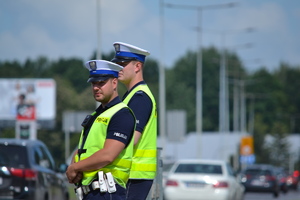 Dwóch poloicjantów ruchu drogowego stoi przy drodze, nadzoruje ruch. W tle ruch uliczny, słoneczna, letnie pogoda